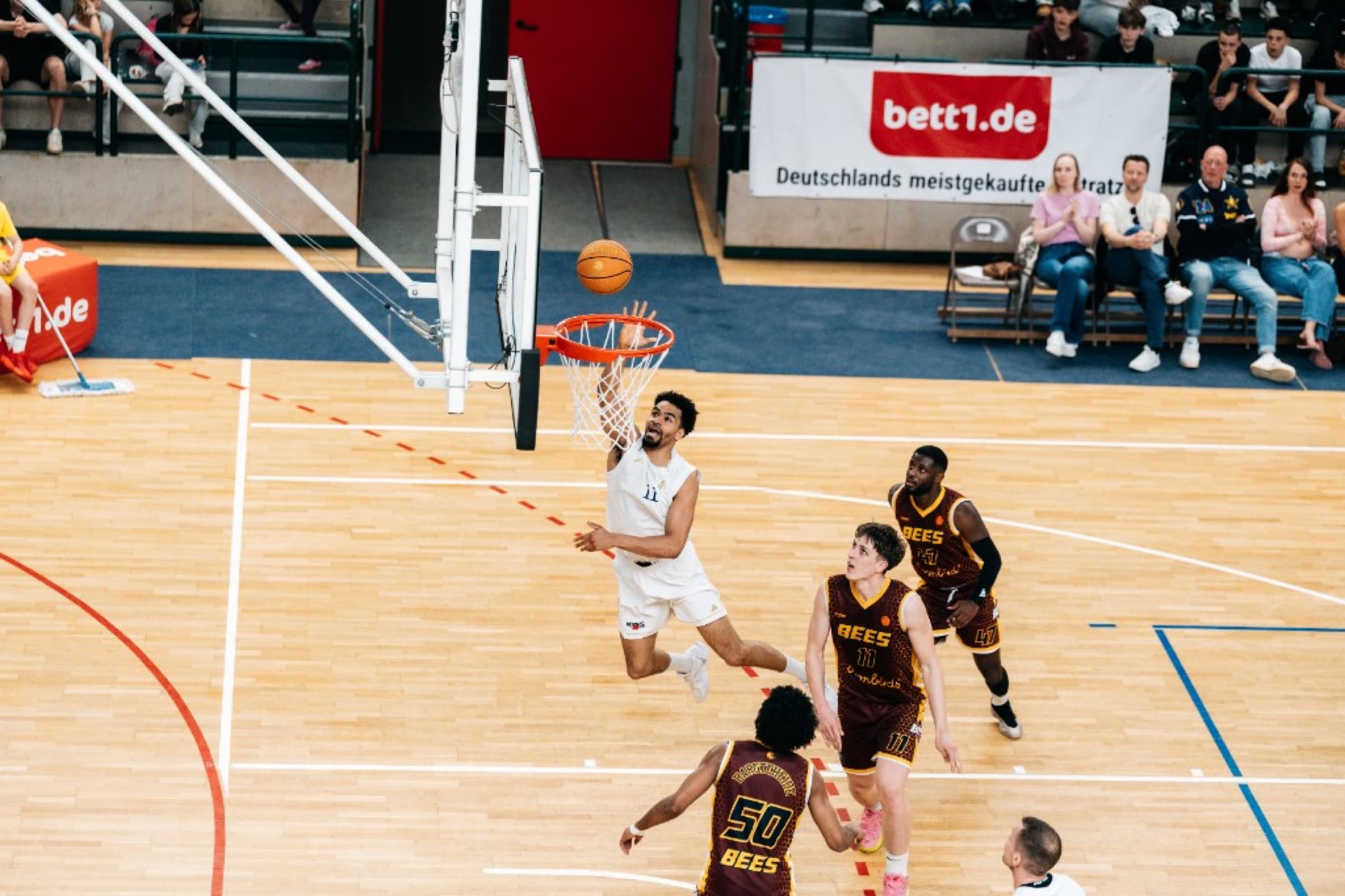 Spieler in Weiß im Sprung zum Layup über Bees-Verteidigern in maroon-gold