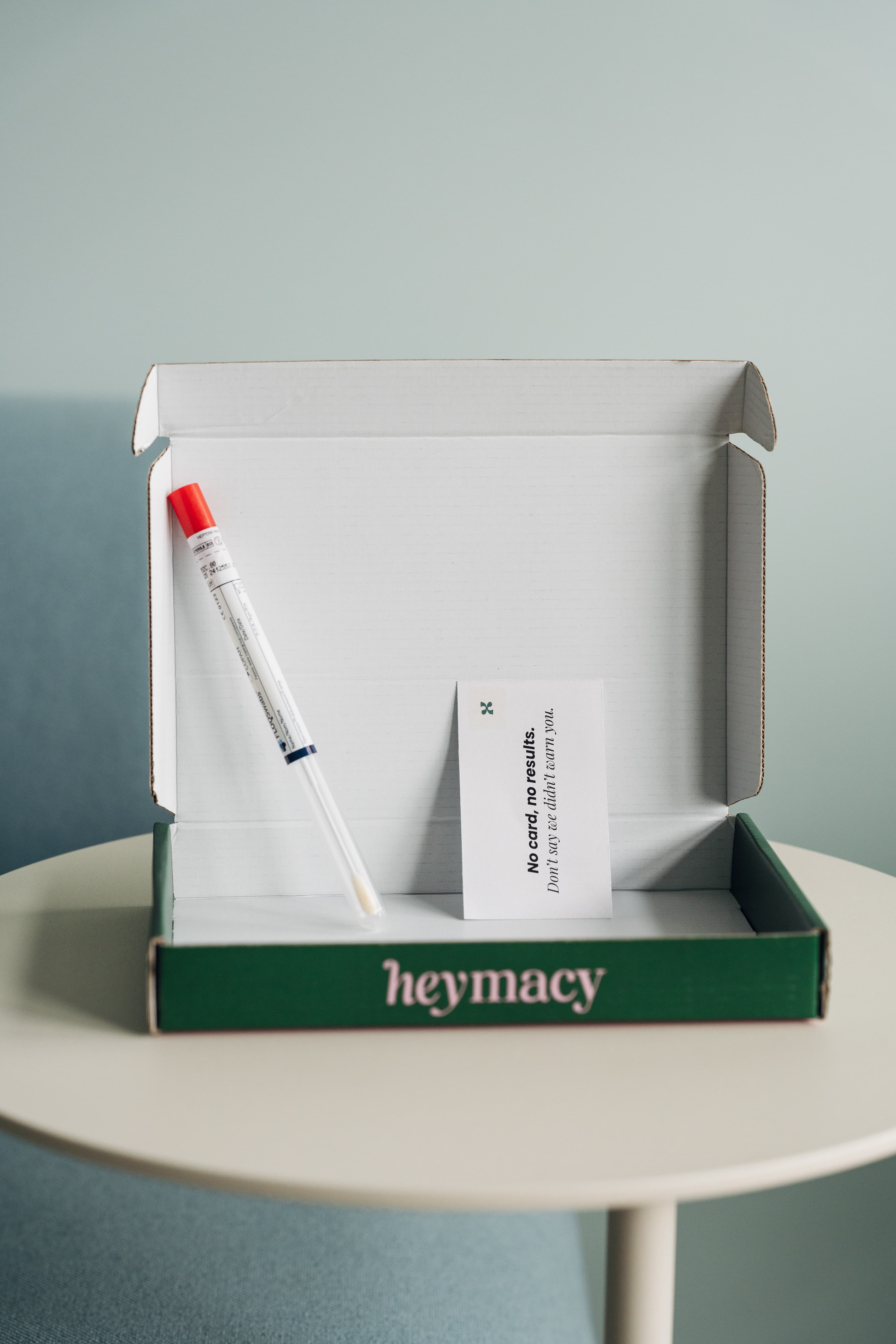 Geöffnete heymacy-Box mit Abstrich-Röhrchen und rotem Deckel und Warnkarte auf dem Tisch