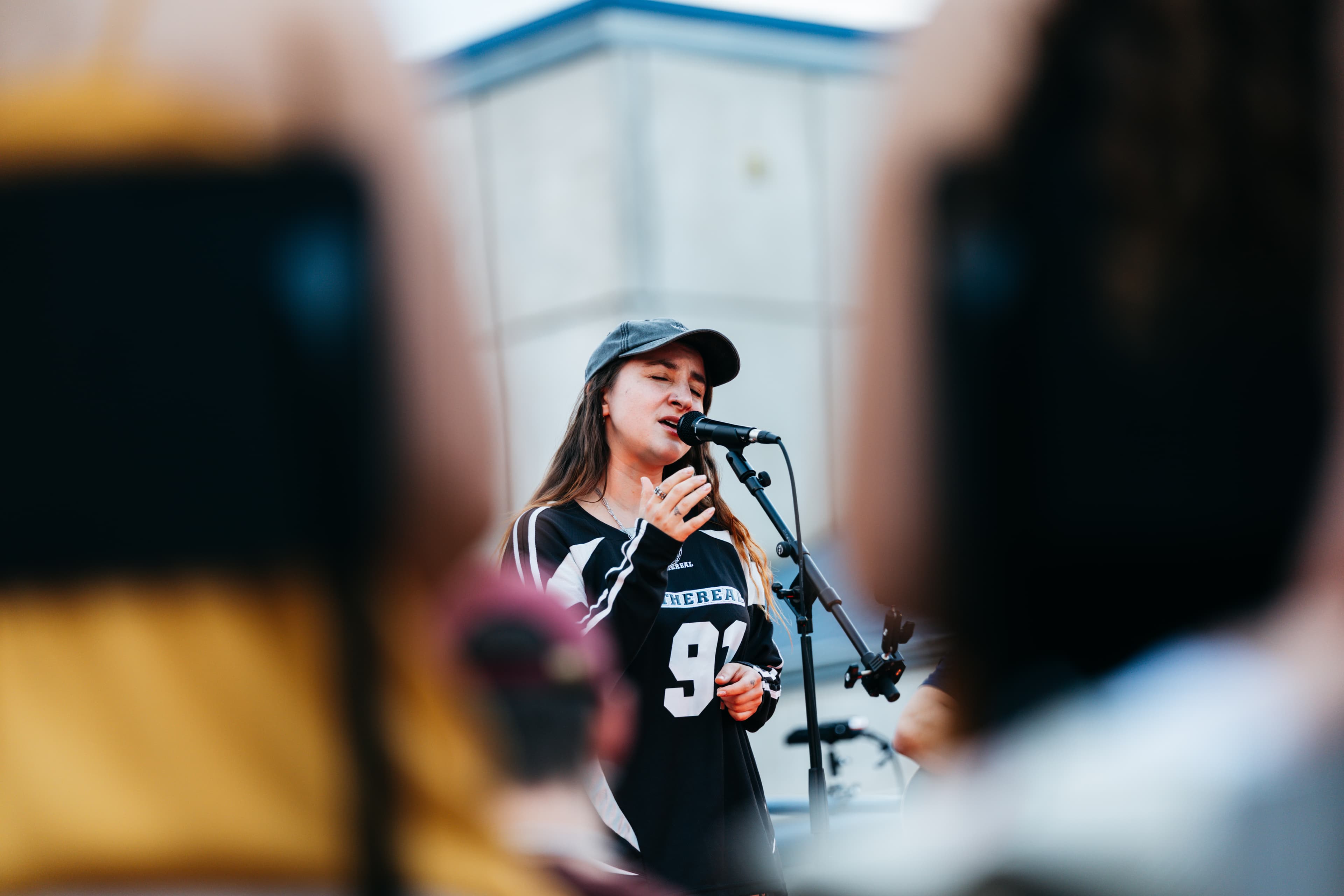 Sängerin mit schwarzem Cap und Jersey 91 singt ins Mikrofon, Publikum unscharf im Vordergrund
