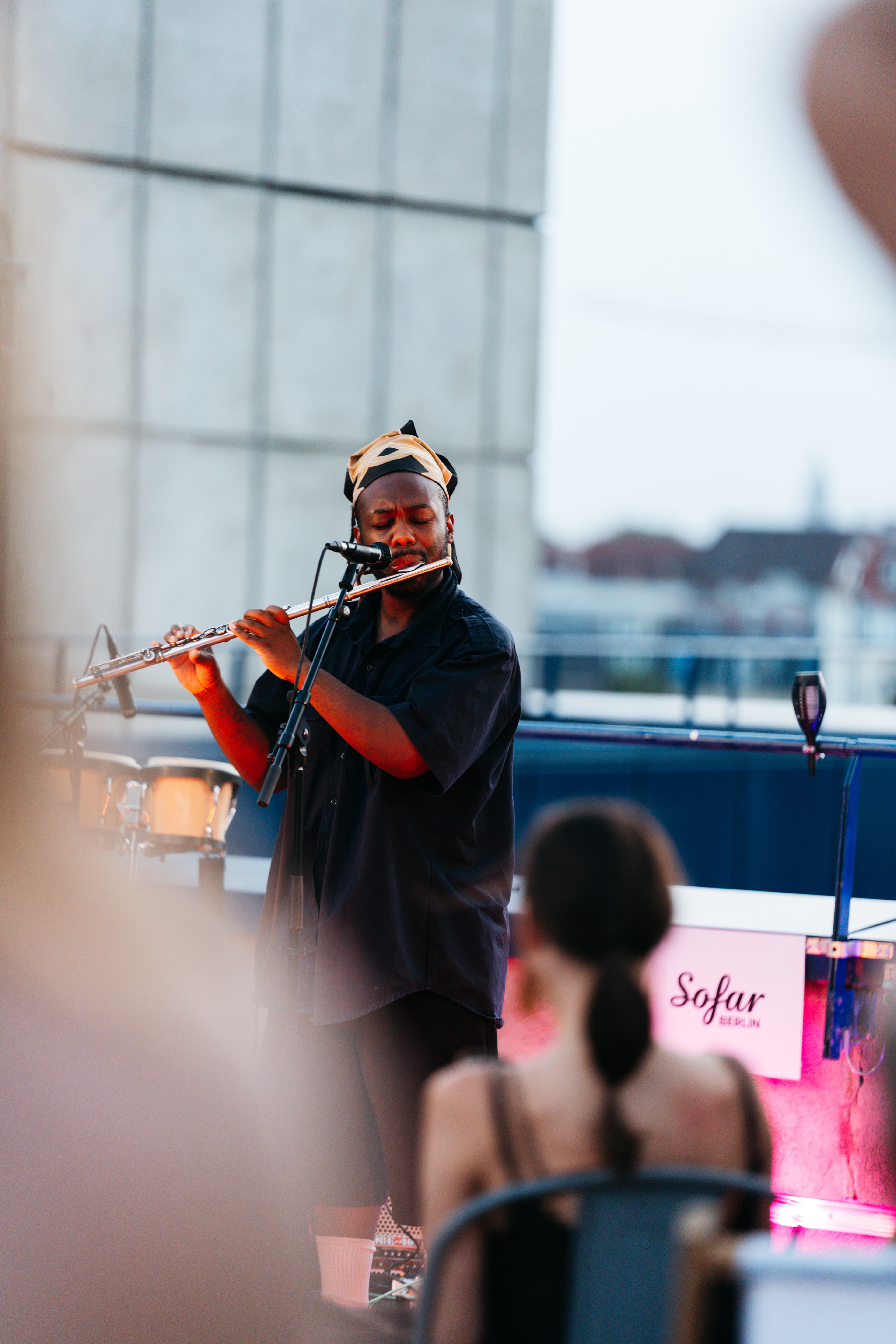 Musiker mit Flöte und Kopftuch bei Sofar Berlin auf dem Dach, warmes Bühnenlicht, Publikum unscharf